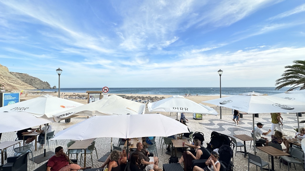 Mesas e chapéus de sol com o mar e a falésia da Luz ao fundo